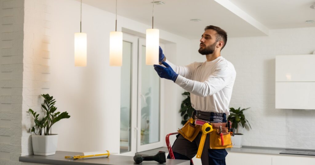 An electrician wearing a tool belt installs modern pendant light fixtures above a kitchen island in a bright home.