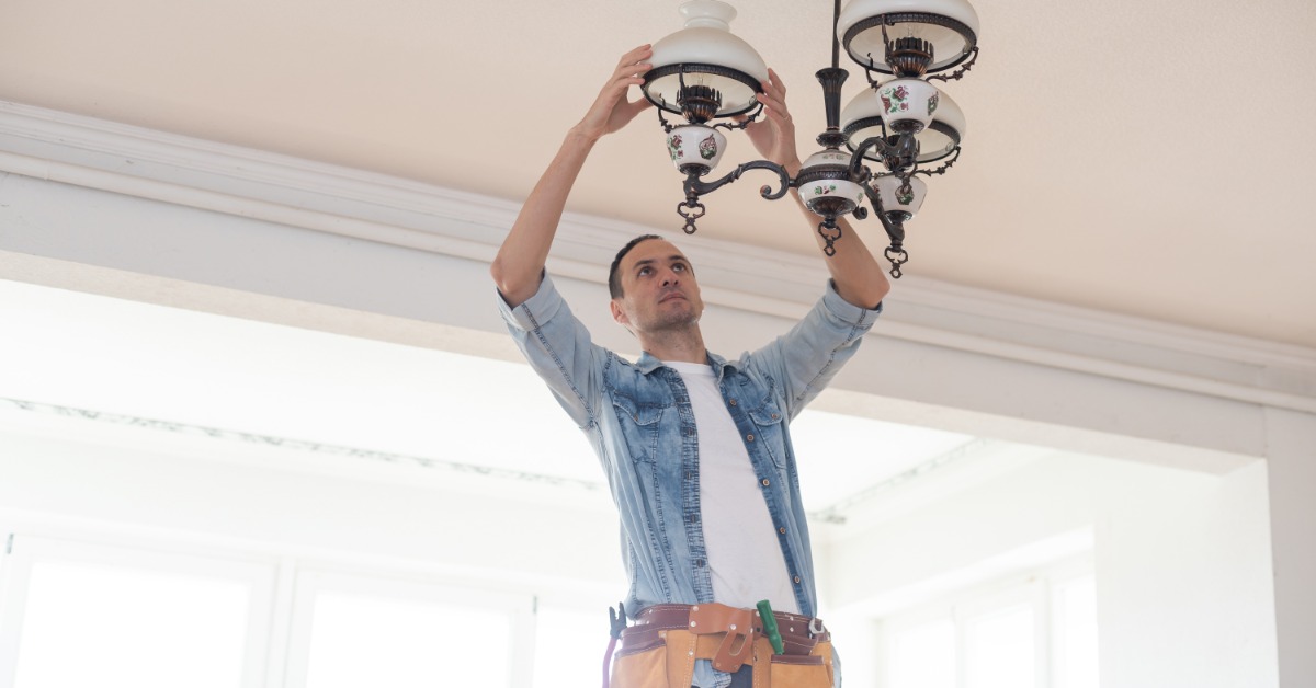 A male electrician with a brown tool belt installs a vintage chandelier with glass shades on a ceiling.