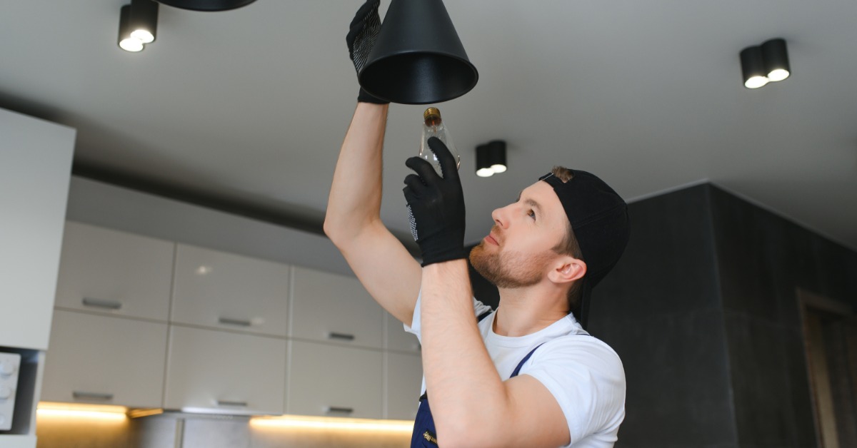 An electrician in blue overalls installs a black pendant light fixture on the ceiling in a modern kitchen.