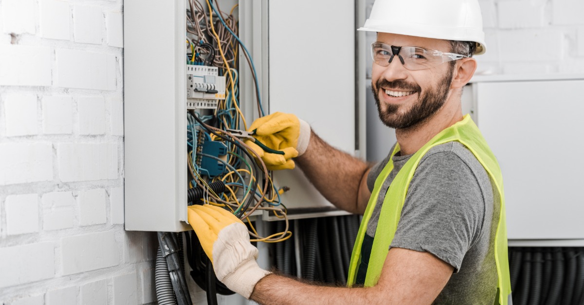 A male electrician in a hard hat and safety glasses works on an open electrical panel mounted on a white brick wall.