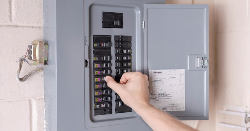 A person adjusting a circuit breaker inside an open gray electrical panel mounted on a wall with labeled switches.