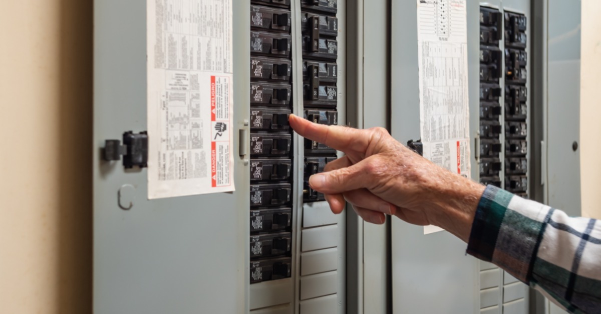 A person pressing a circuit breaker switch on a gray electrical panel with labeled breakers and multiple panels.