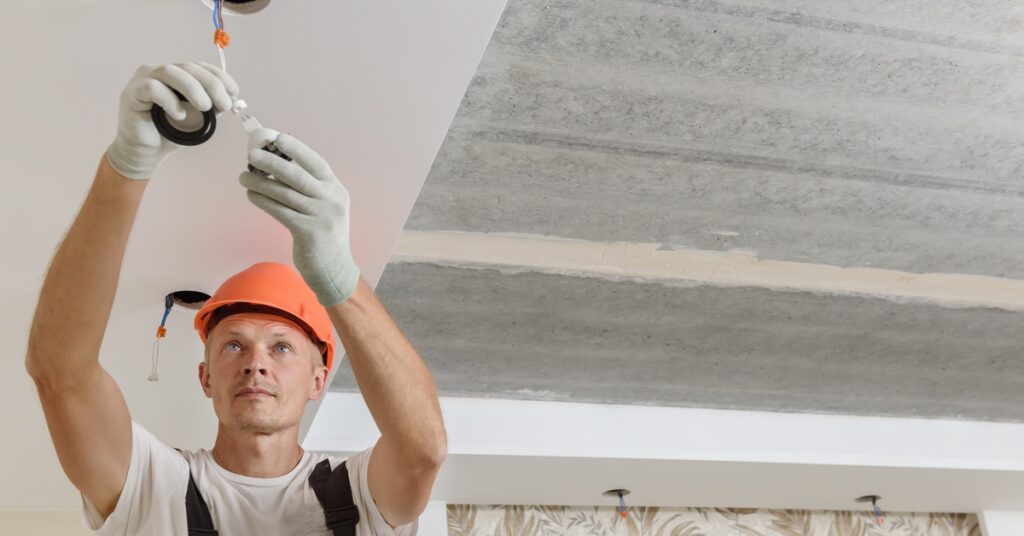 An electrician in an orange hard hat installs ceiling light wiring beneath an unfinished concrete ceiling.