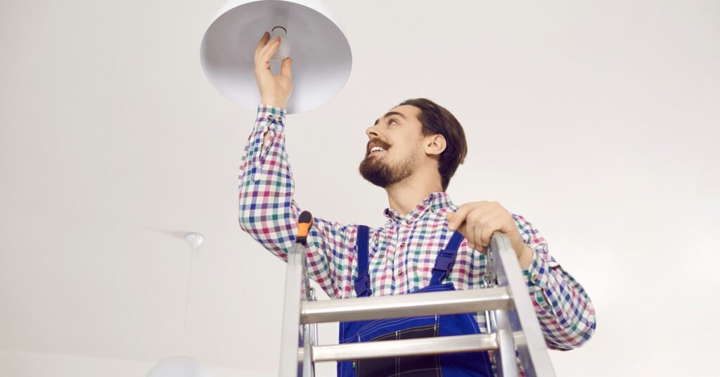 A man in overalls stands on a ladder installing a ceiling light fixture, reaching up with one hand while smiling.