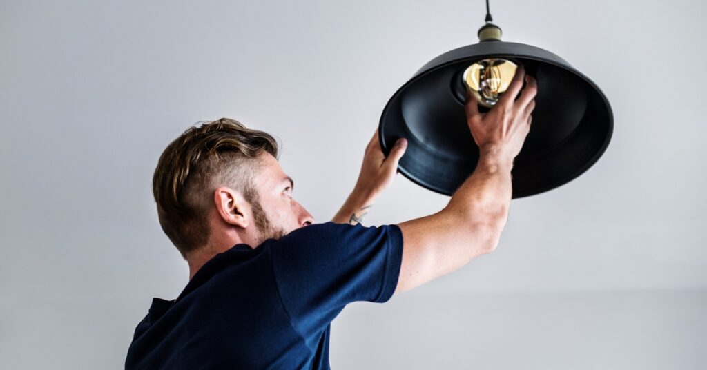 A man installs a light bulb in a black hanging ceiling fixture, reaching up with both hands against a plain white background.