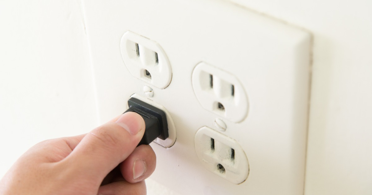 An individual plugging a black power cord into a white wall outlet with four sockets on a clean interior wall.