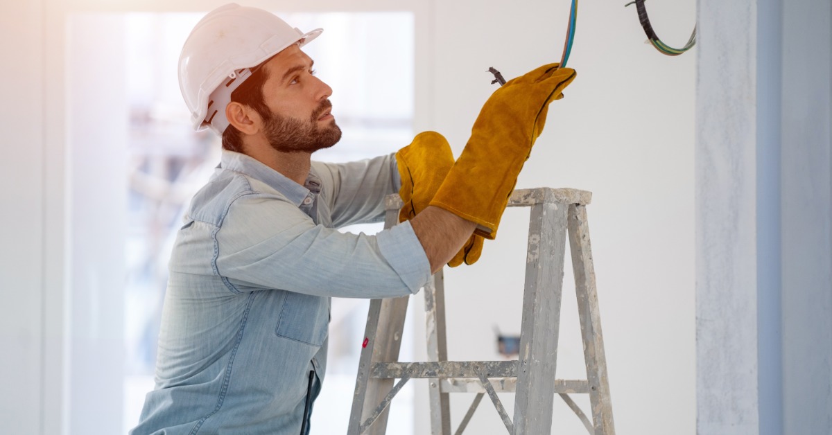 An electrician wearing safety gloves and a hard hat repairs exposed ceiling wiring while standing on a ladder indoors.