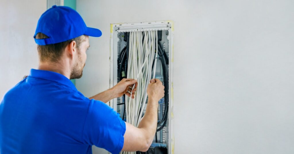 An electrician in a blue uniform organizing wiring inside an open electrical panel mounted on a white wall.