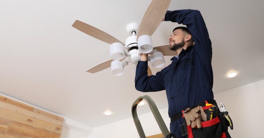An electrician installs a modern ceiling fan while standing on a ladder in a bright room with recessed lighting.