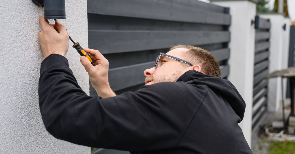 An electrician adjusts an outdoor wall light fixture with a screwdriver beside a modern black residential fence.