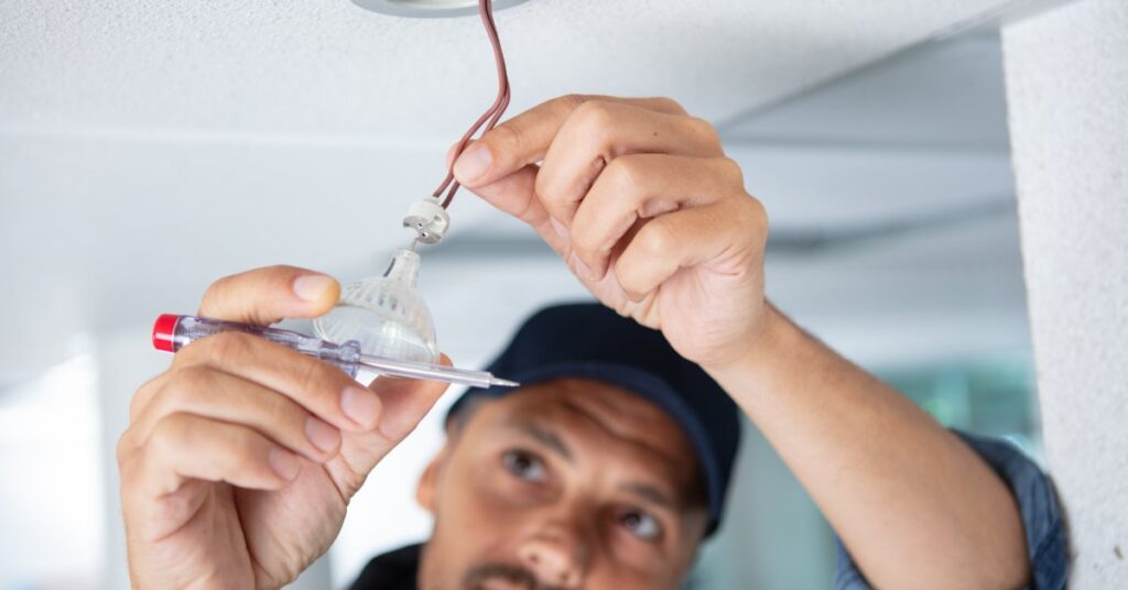 An electrician connects wiring to a recessed ceiling light fixture while holding a voltage tester during installation.