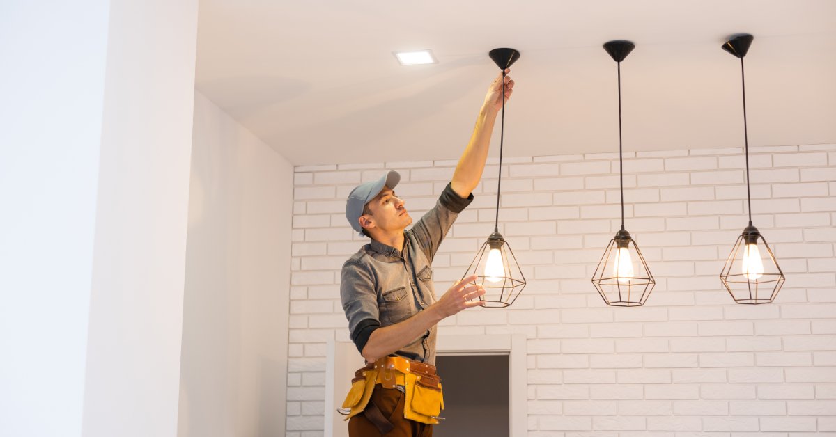 An electrician adjusts a hanging pendant light fixture in a modern room with three illuminated ceiling lights.
