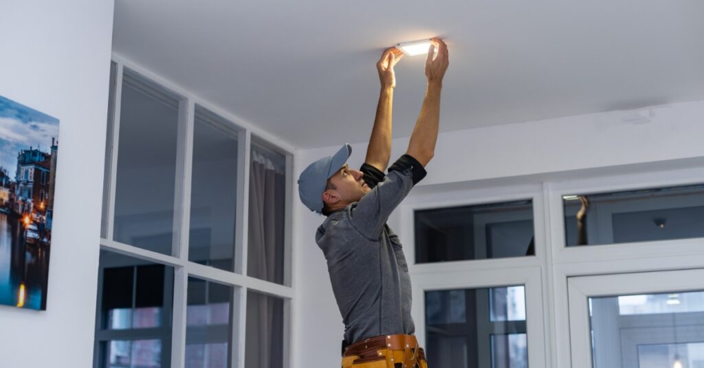 An electrician reaches overhead to install a ceiling light fixture in a bright room with large windows and white walls.