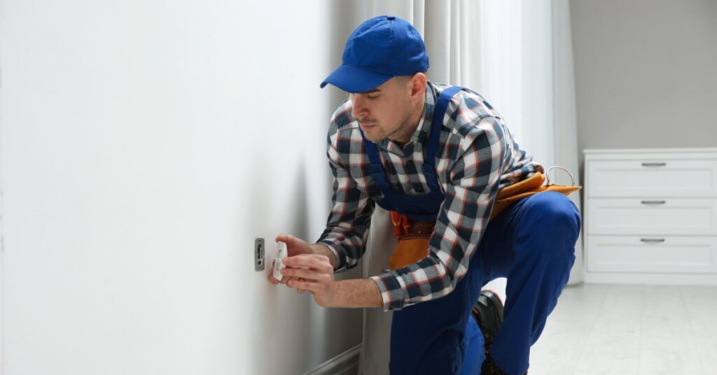 An electrician kneels beside a wall installing an electrical outlet while tools rest on the floor in a bright room.