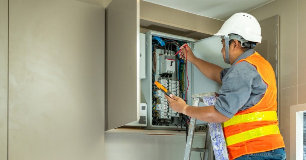 Electrician in a hard hat testing wiring inside an open electrical panel while standing on a ladder during service.