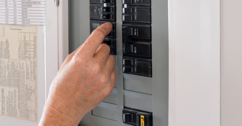 Hand pressing a circuit breaker inside a residential electrical panel with labeled switches mounted on a white wall.