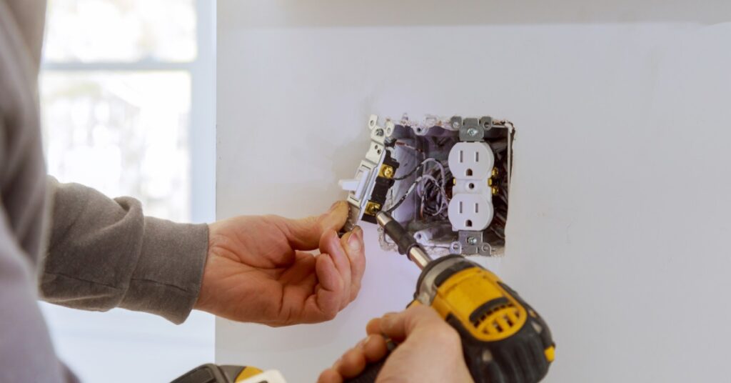Electrician uses a drill on an exposed wall box containing a light switch, electrical outlet, and visible wiring.