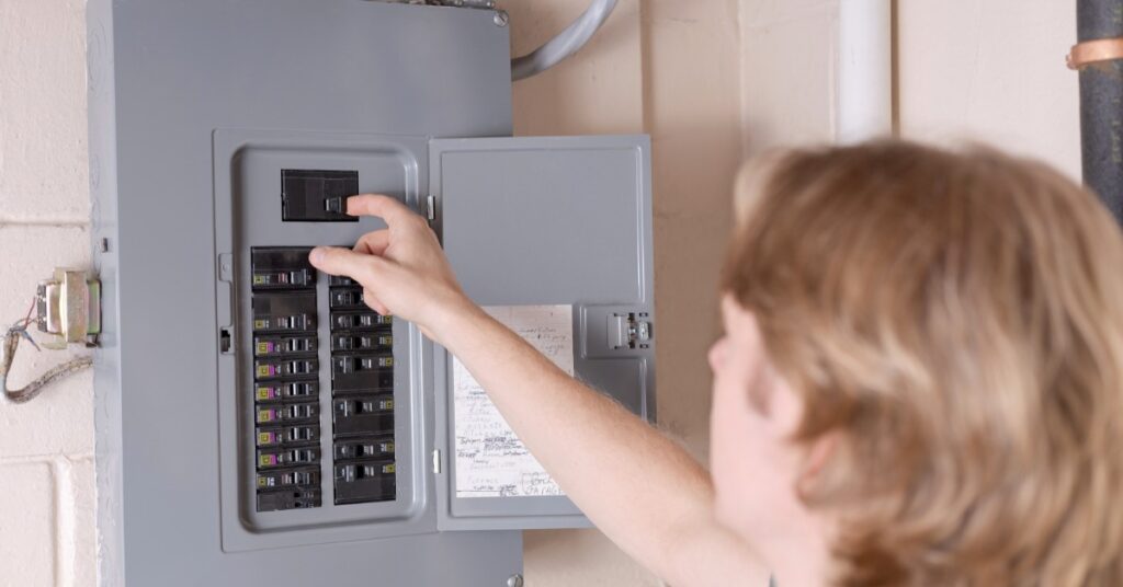 Person reaching to switch a circuit breaker inside an open gray electrical panel mounted on a white wall.