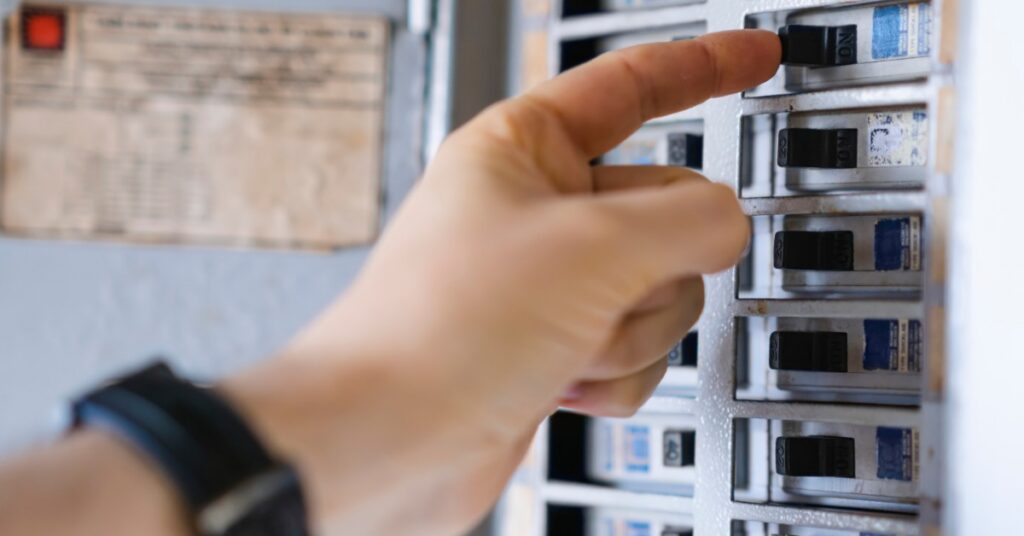 Hand pressing a black circuit breaker switch inside an electrical panel filled with rows of breakers.