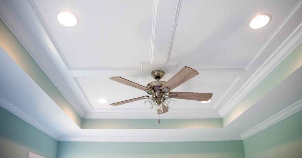 White tray ceiling with recessed lights and a central wooden ceiling fan above pale green walls and decorative molding.