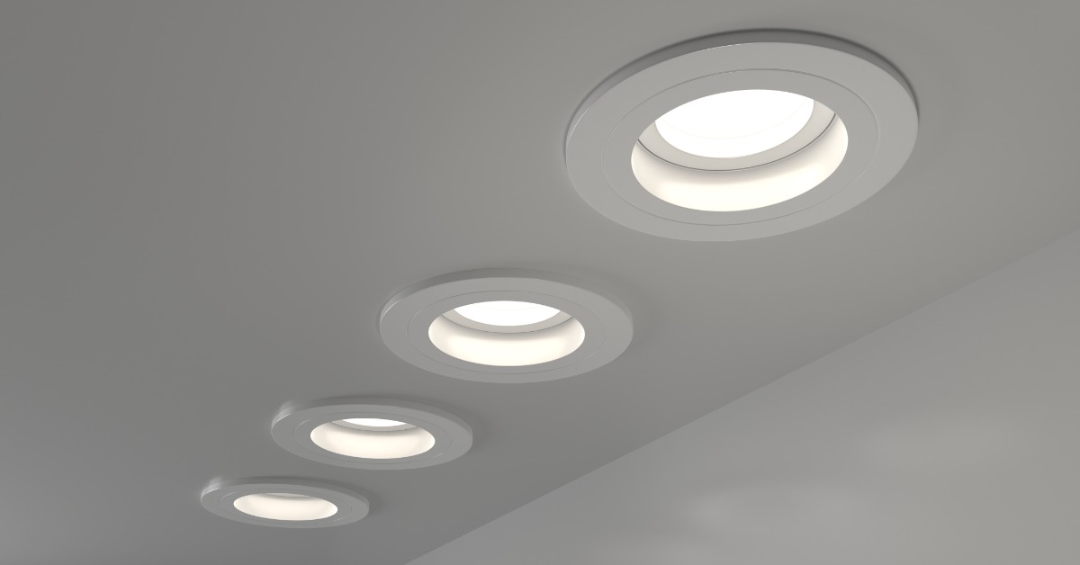Four illuminated recessed ceiling lights with circular white trim arranged diagonally across a smooth light gray ceiling.